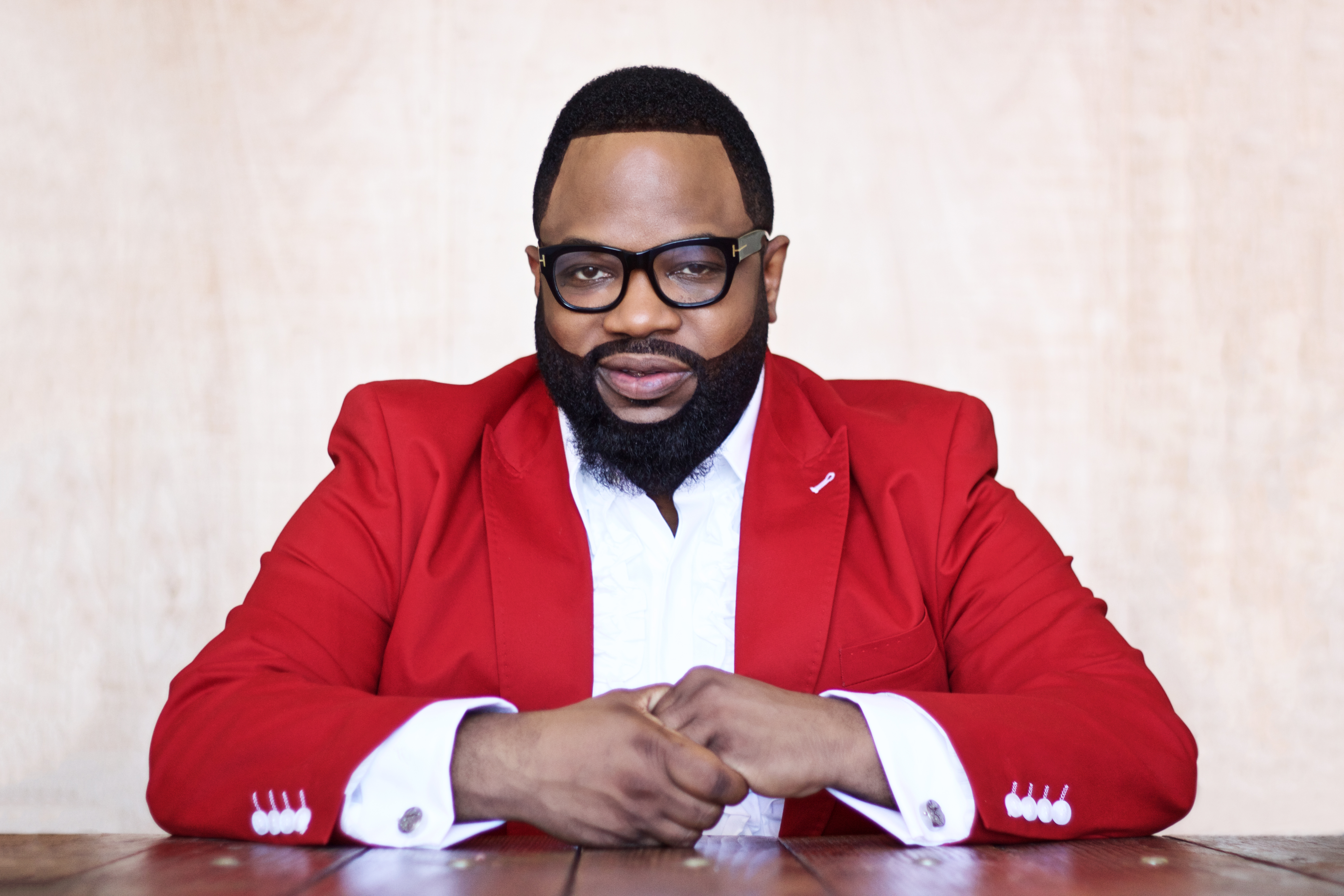A man wearing a red suit and glasses, with a full beard, sitting at a table and looking directly at the camera.