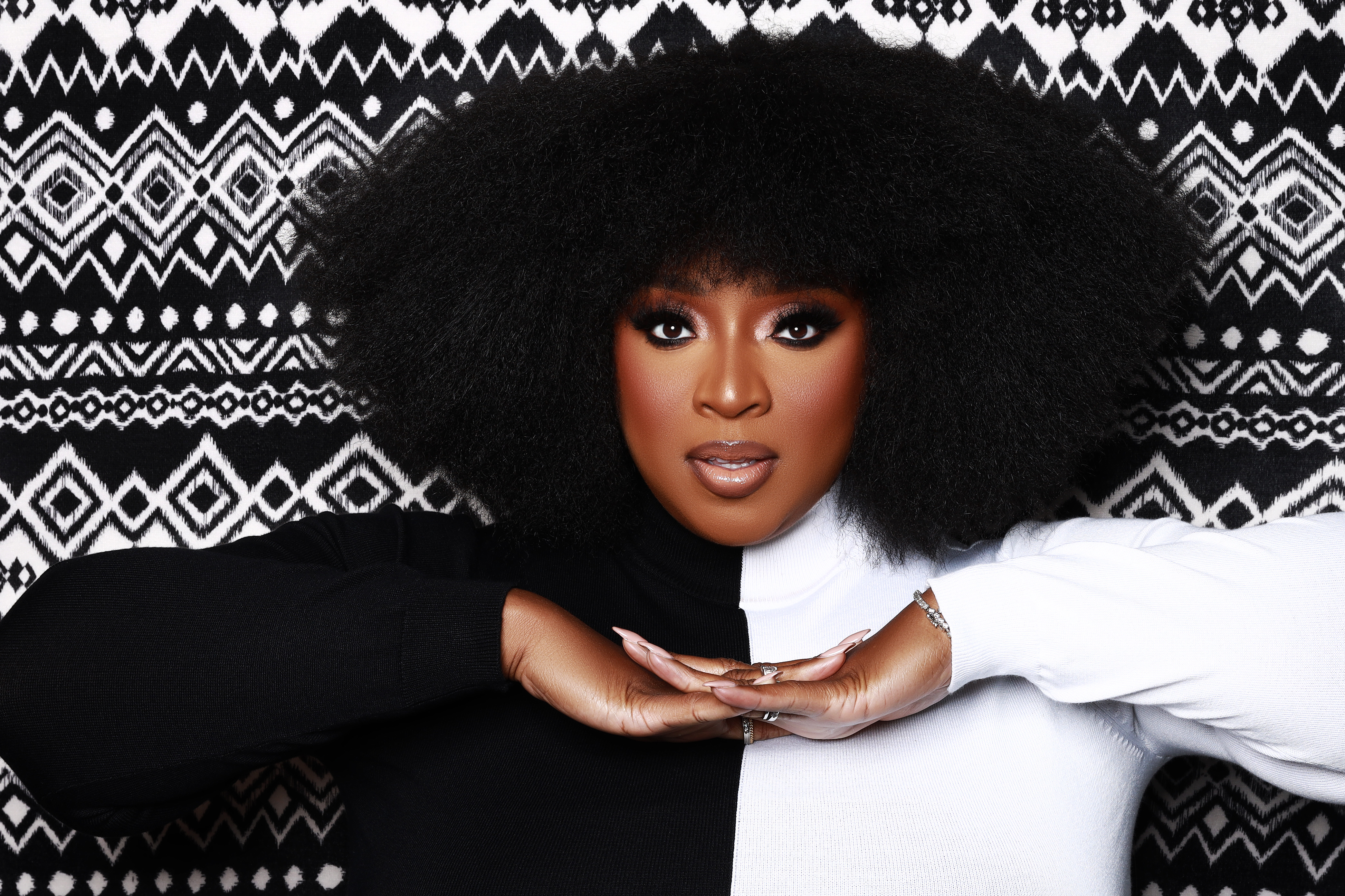 A close-up portrait of a Black woman with a large, curly afro hairstyle against a black and white patterned background.