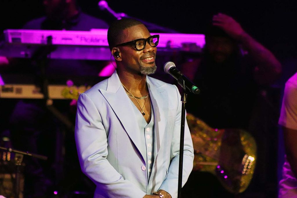 A man with a beard and glasses speaking into a microphone on a stage with colorful lighting.