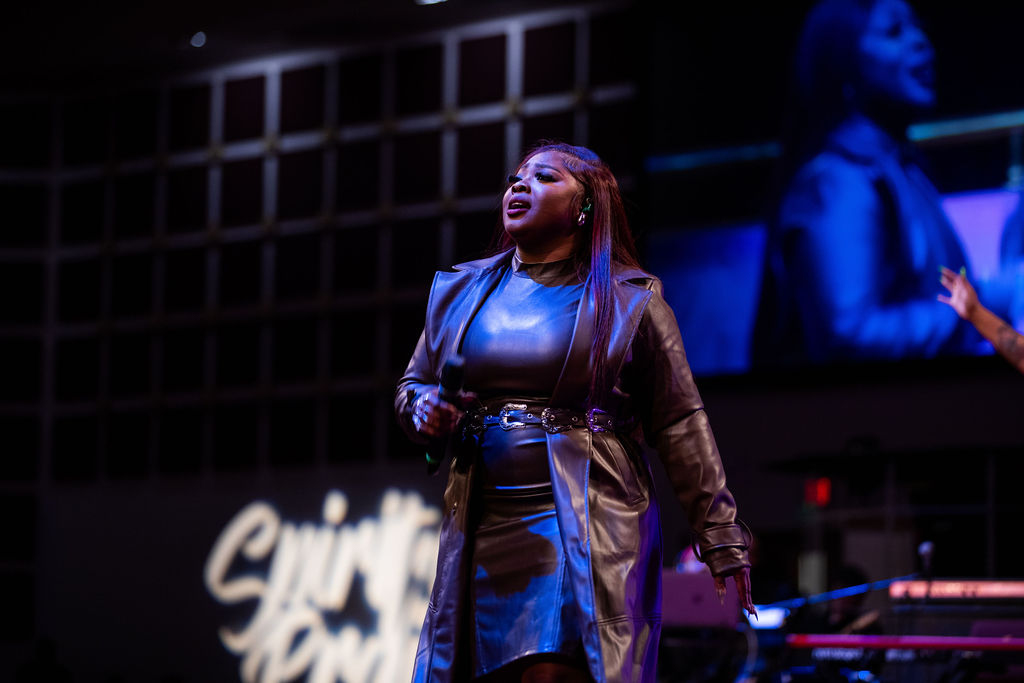 A woman in a leather jacket and pants stands on a stage, singing into a microphone with a dramatic expression.