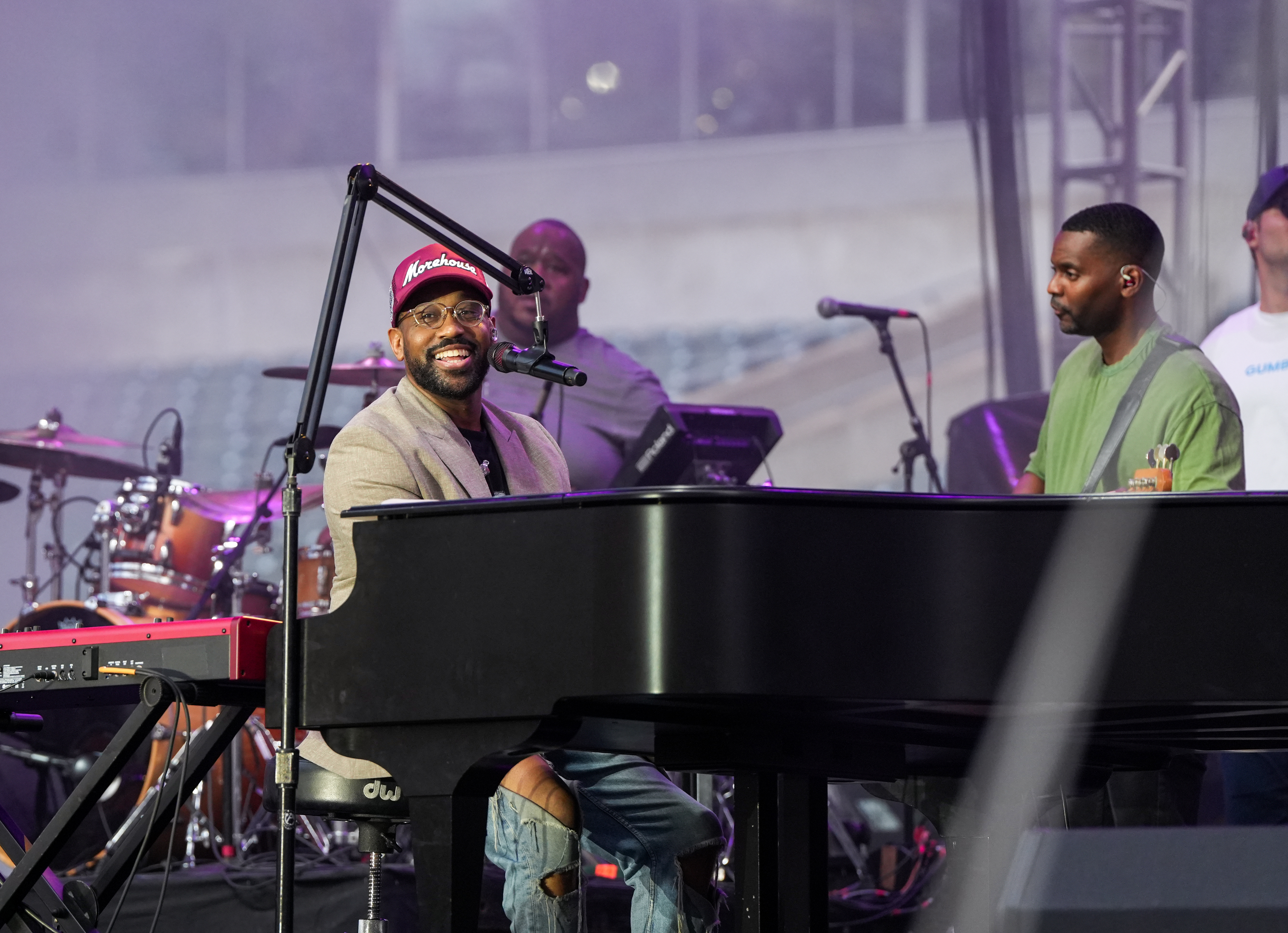 A smiling man playing a piano on stage, wearing a Morehouse cap, with a drummer and another musician visible behind him.