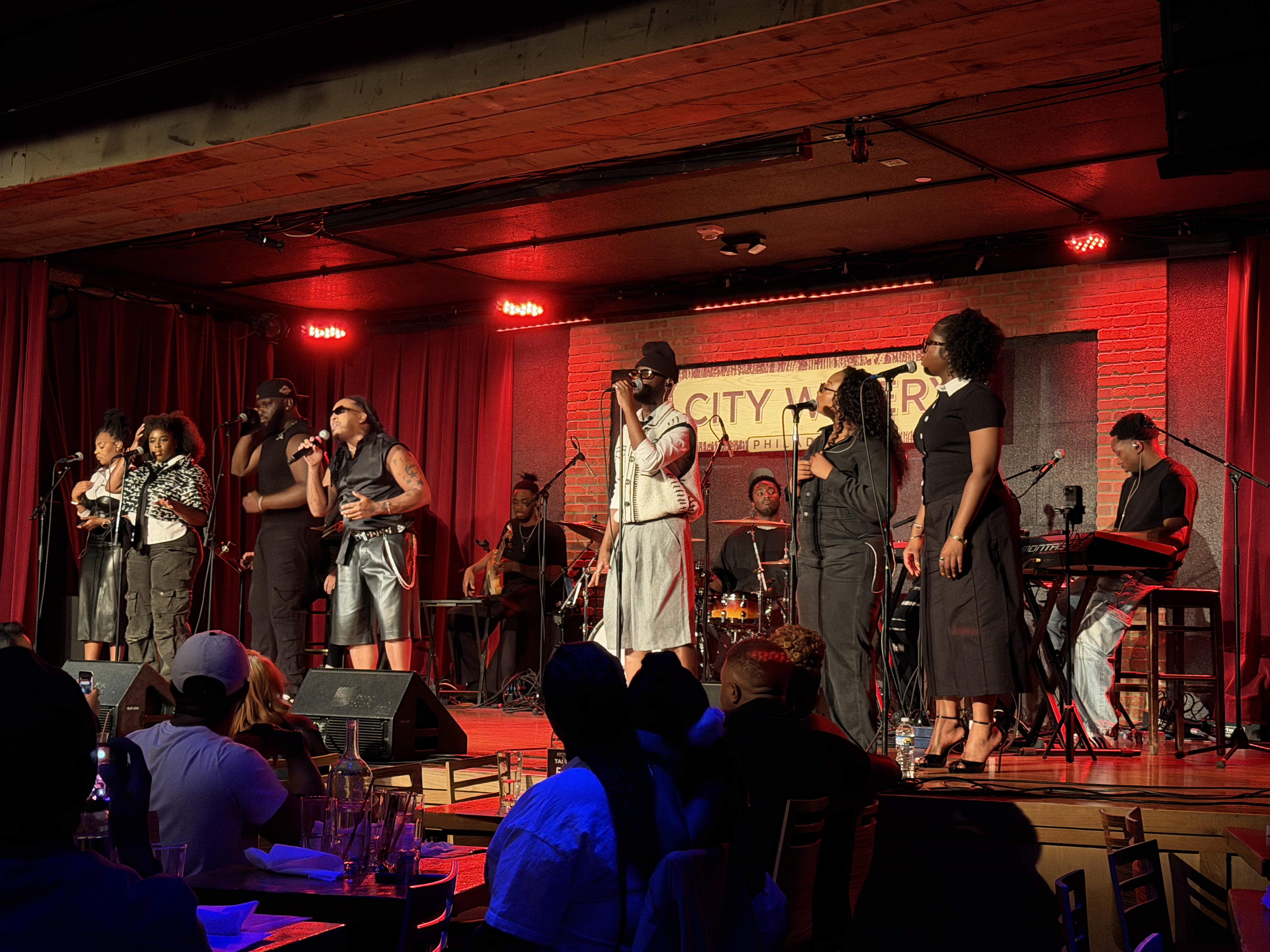 A live music performance on a stage with a red curtain backdrop, featuring a band and vocalists singing and playing instruments in front of an audience.