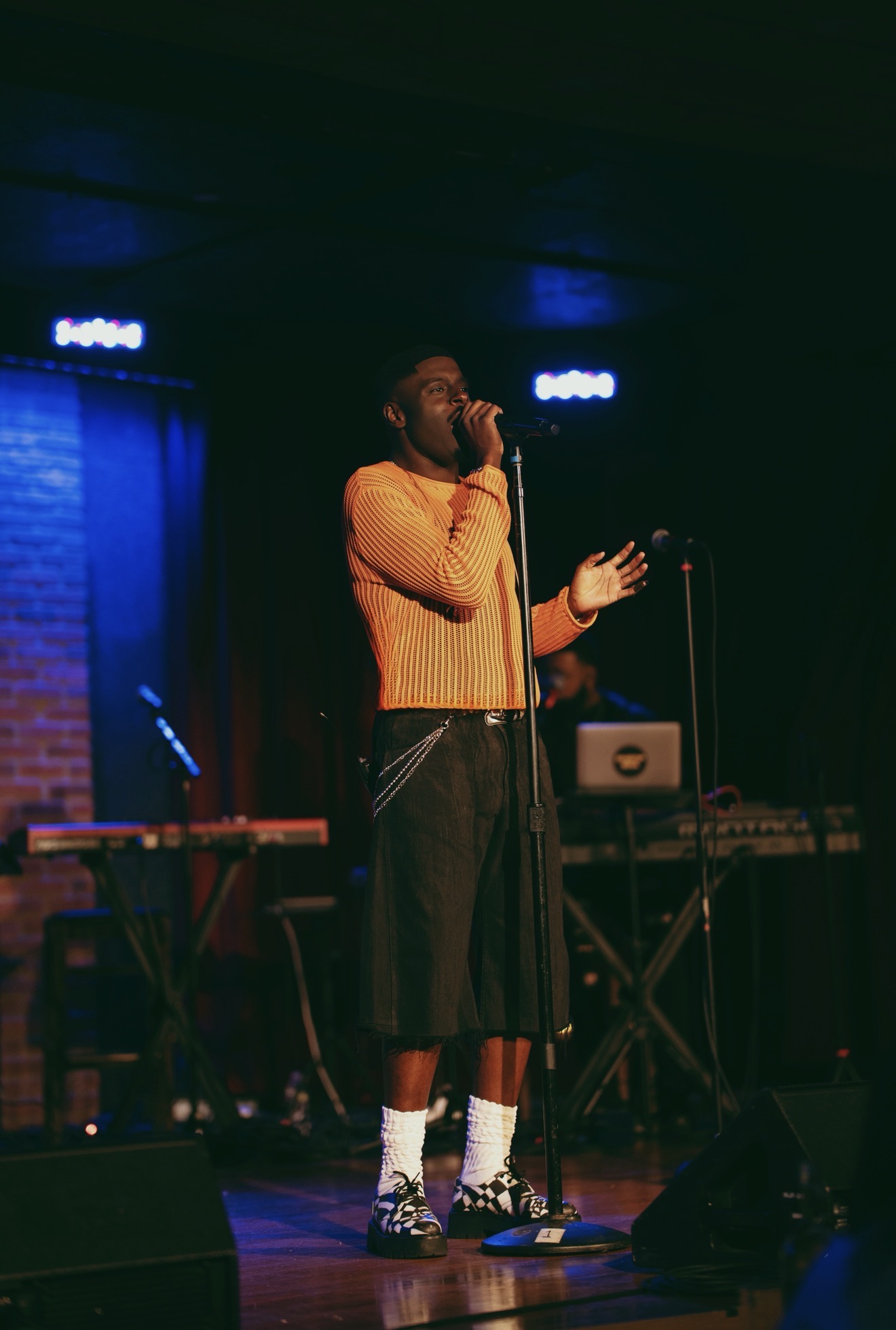 A man in an orange sweater performing on stage, with musical equipment and lighting visible in the background.