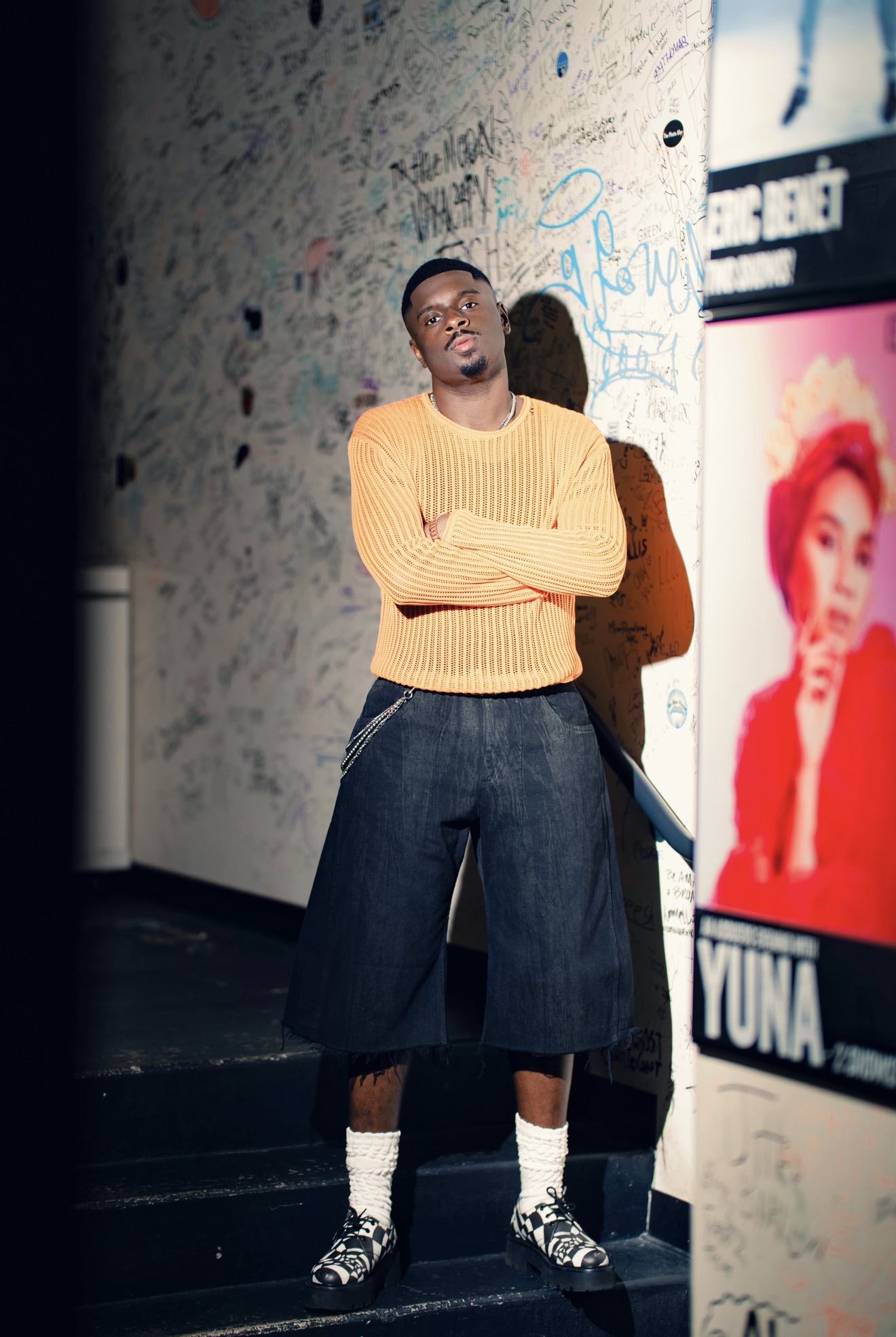 A young man in a yellow sweater stands with his arms crossed in front of a graffiti-covered wall, with a large "YUNA" poster visible.