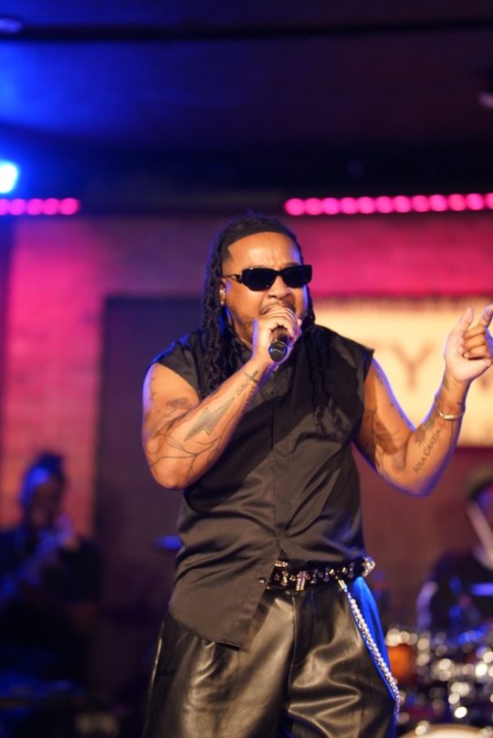 A man wearing sunglasses and a black outfit performing on stage with a microphone.